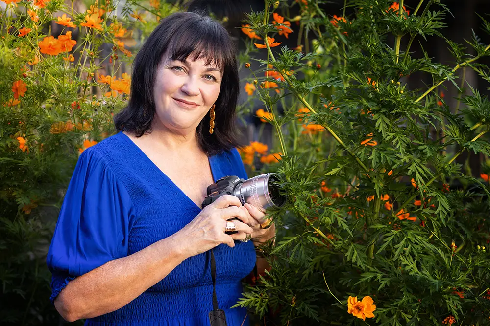 Photographs of Cheryl Eagers supplied as reference, with her DSLR among cosmos flowers, and the portrait the character was drawn from (1 of 2)